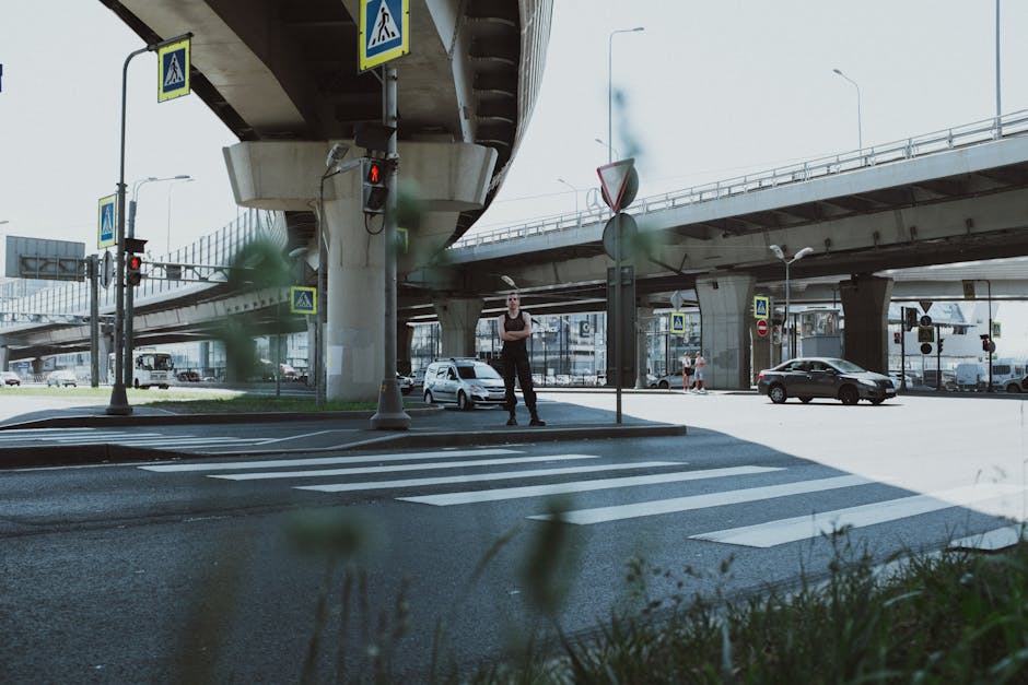 A pedestrian waiting at a crosswalk under an urban overpass with cars passing.